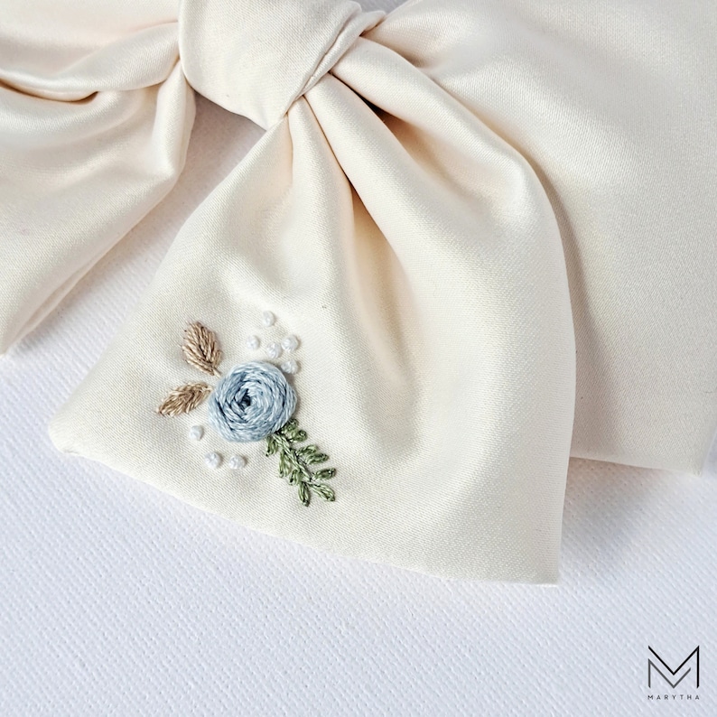 May include: A cream-colored satin bow with a delicate embroidered floral design. The design features a light blue flower with green leaves and white pearl-like accents.