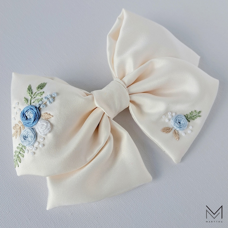 May include: A large ivory satin bow with blue and white embroidered flowers. The bow is perfect for adding a touch of elegance to any outfit.