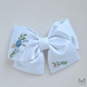 May include: A white satin bow with embroidered blue and green floral designs. The bow is tied in a classic shape with a center loop.