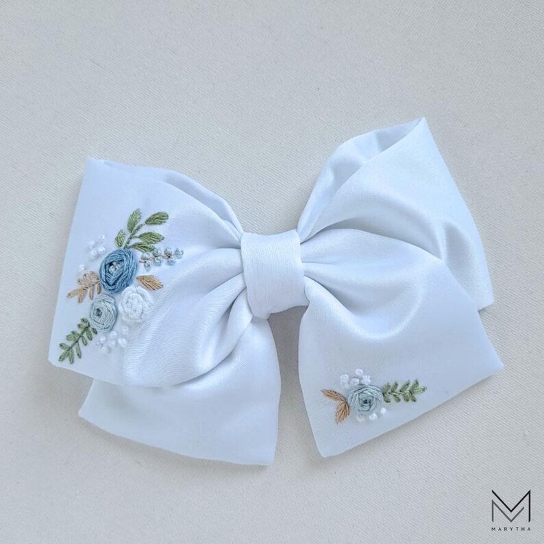 May include: A white satin bow with embroidered blue and green floral designs. The bow is tied in a classic shape with a center loop.