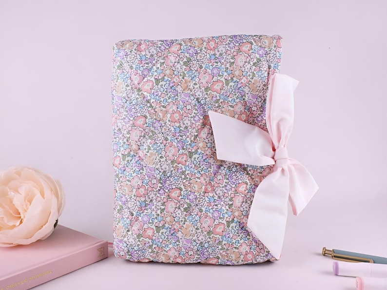 May include: A rectangular quilted fabric book cover featuring a dense floral pattern in shades of pink, blue, and green. The cover is secured with a large, light pink satin ribbon tied into a bow on the right side, adding a soft texture.