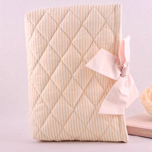 May include: A quilted book cover featuring a light beige and white vertical striped pattern with a diamond-stitched texture. The cover is accented with a large, soft pink fabric bow attached to the right side, providing a decorative closure detail.