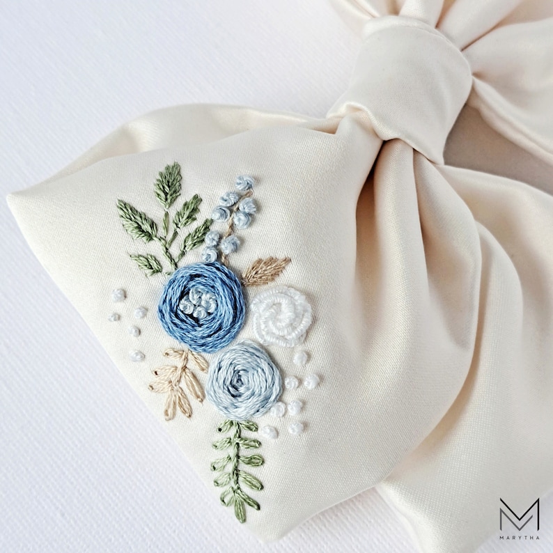 May include: A white satin bow with blue and white embroidered flowers. The flowers are in a cluster and include leaves and small white dots.