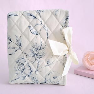 May include: A quilted fabric book cover featuring a cream-colored base with a dark blue floral pattern. The cover is secured with a cream-colored ribbon tied in a bow on the right side. The quilted texture consists of a diamond-shaped stitching pattern.