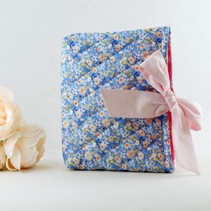 May include: A rectangular quilted fabric cover featuring a blue floral pattern with small pink and white flowers. The cover is secured with a light pink satin ribbon tied in a bow on the right side. The interior lining is a solid bright pink fabric.