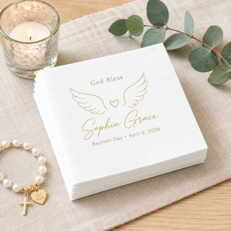 Op de afbeelding: Een stapel vierkante witte papieren servetten met goudkleurige foliebedrukking. Het ontwerp bevat engelvleugels, een klein hartje en de gedrukte tekst God Bless, Sophia Grace, Baptism Day en de datum 6 april 2028 in een sierlijk lettertype.