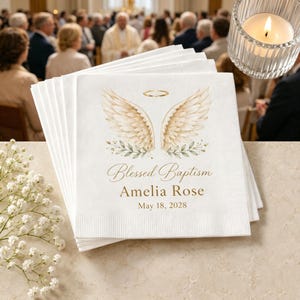 Op de afbeelding: Een stapel witte papieren servetten met een illustratie van een gouden halo en beige engelenvleugels boven groene bladeren. De tekst Blessed Baptism, Amelia Rose en May 18, 2028 staat in een gouden lettertype op het voorste servet.