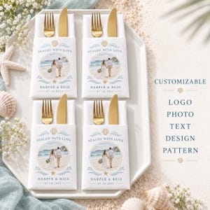 May include: Four white paper cutlery pouches, each holding a gold fork and knife. The pouches feature a circular photo of a couple on a beach, the text "SEALED WITH LOVE," "HARPER & REID," and "07.18.2031," with blue wave and gold seashell accents.