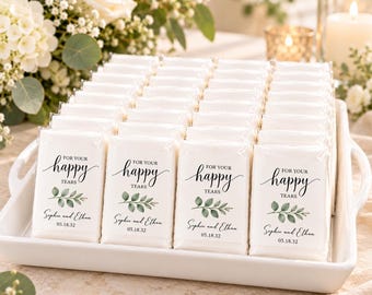 Op de afbeelding: Een verzameling rechthoekige zakdoekjes in een witte schaal. Elk pakje is voorzien van een wit label met de tekst "FOR YOUR happy TEARS", een illustratie van een groen blad, de namen "Sophie and Ethan" en de datum "05.18.32".