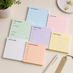 May include: A selection of pastel-coloured sticky note pads for planning and organisation. The pads feature sections for top priorities, daily tasks, goal tracking, weekly goals, habit tracking, notes, and weekly plans. A pen and bookmark are also present.