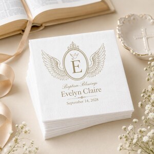 Op de afbeelding: Een stapel witte papieren servetten met een goudkleurige opdruk van een ovaal kader met vleugels, een kleine duif en de letter E. Onder de afbeelding staat de tekst Baptism Blessings, Evelyn Claire en September 14, 2028.
