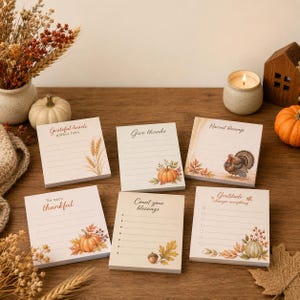 May include: Six notepads with autumn designs. Each notepad has a different phrase, including "Grateful hearts gather here" and "Give thanks." The notepads are decorated with pumpkins, leaves, and a turkey, evoking a harvest theme.