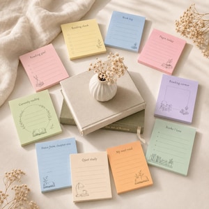 Op de afbeelding: Een verzameling kleurrijke notitieblaadjes met verschillende titels en illustraties over lezen. De blaadjes zijn gerangschikt rond een kleine witte vaas met gedroogde bloemen en verschillende boeken. De blaadjes zijn in pastelkleuren.