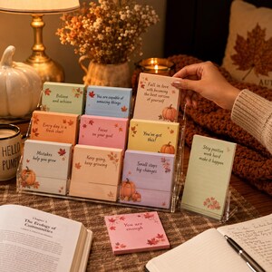 Könnte beinhalten: Ein klarer Schreibtisch-Organizer aus Acryl mit bunten Haftnotizen mit inspirierenden Zitaten und herbstlichen Illustrationen. Die Notizen zeigen Kürbisse und Herbstblätter. Eine Hand hält eine der Notizen. Ein Buch und eine Tasse sind im Hintergrund.