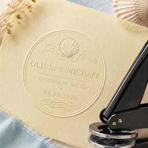 May include: A cream-colored paper embossed with a circular design featuring a seashell icon, the text OLIVIA & MICHAEL, TOGETHER BY THE SEA, and the date 07.18.2026. A black metal embosser tool is positioned on the right side of the paper.