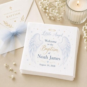 Op de afbeelding: Een stapel witte papieren servetten met blauwe engelvleugels, een gouden halo en de tekst Little Angel Welcome to the Baptism of Noah James August 10, 2028, versierd met kleine blauwe en gouden sterren en een klein blauw hartje.