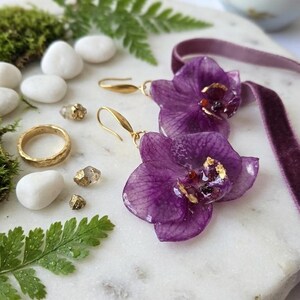 Puede incluir: Un par de pendientes de orquídeas moradas con ganchos dorados, un anillo dorado texturizado y una cinta de terciopelo morado. Las joyas están dispuestas junto a pequeñas piedras blancas, hojas de helecho verdes y fragmentos minerales dorados.