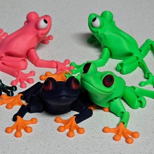 May include: Four articulated frog figurines in different colors: pink, green, black, and green. The frogs have large eyes and are posed in a sitting position.
