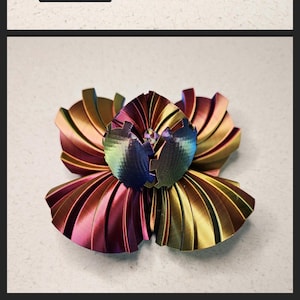 May include: A multi-coloured, iridescent, 3D printed sphere with a flower-like design. The sphere is made up of multiple interlocking pieces that can be rotated and rearranged.