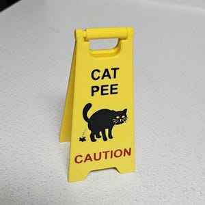 Pode incluir: Um sinal de advertência amarelo com uma silhueta de gato preto e o texto "CAT PEE CAUTION".
