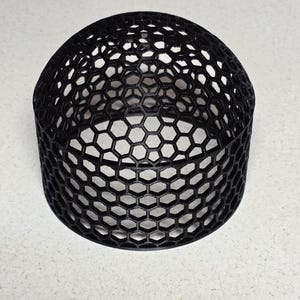 Puede incluir: Un objeto negro en forma de cúpula con un patrón de panal. El objeto es cilíndrico con una parte superior redondeada, y el diseño abierto revela la superficie debajo. El objeto está hecho de un material oscuro y se encuentra sobre una superficie clara.