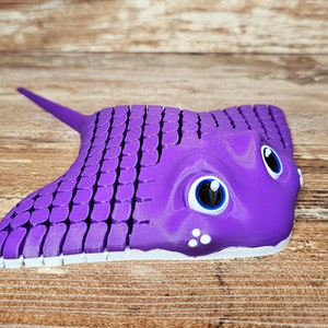 May include: A purple 3D printed stingray toy with white accents and blue eyes. The stingray has a textured surface and a long tail.