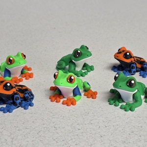 May include: A collection of six colorful frog figurines. The frogs feature vibrant colors including green, orange, blue, and white. Each frog has large, expressive eyes and detailed features, perfect for collectors or as decorative items.