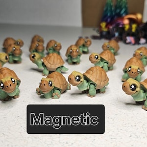 May include: A collection of small, green and brown turtle figurines with magnetic features. The turtles have cute faces and are arranged in a row on a white surface. The word "Magnetic" is written in white letters on a black background below the turtles.
