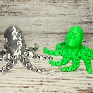 Może przedstawiać: Dwie 3D-wydrukowane rzeźby ośmiornic z ludzkimi głowami. Jedna jest srebrna, a druga zielona. Rzeźby znajdują się na drewnianej powierzchni.