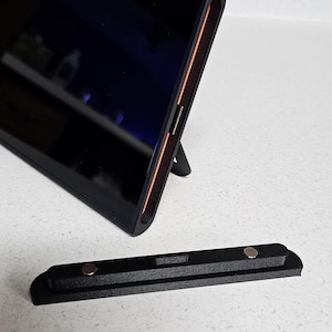 May include: A black tablet with a built-in stand, angled slightly upward. The tablet has a sleek design with a thin profile and a dark screen. A separate black magnetic base is in front of the tablet.