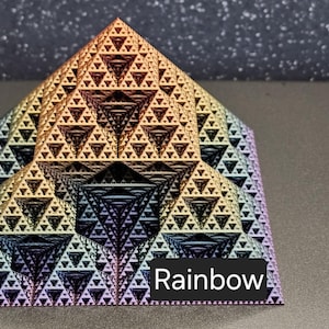 Puede incluir: Una pirámide fractal de color arcoíris hecha de pequeños triángulos. La pirámide está sentada sobre una superficie marrón. La palabra "Rainbow" está impresa debajo de la pirámide.
