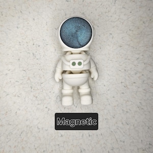 May include: White astronaut figurine with a blue and green swirl pattern on the helmet. The figurine is magnetic and has the word "Magnetic" printed below it.