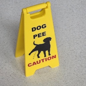 Pode incluir: Um sinal de aviso amarelo com uma pega, com o texto "DOG PEE" acima de uma silhueta preta de um cão a urinar, e "CAUTION" a vermelho por baixo. O sinal está numa superfície cinzenta salpicada.