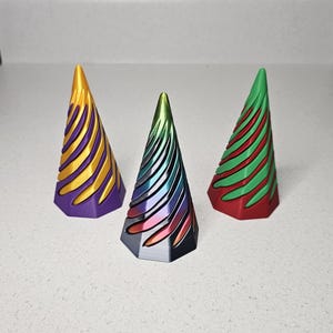May include: Three small, decorative, cone-shaped Christmas trees. The trees are made of plastic and have a metallic finish. The tree on the left is purple with gold stripes, the middle tree is black with rainbow stripes, and the tree on the right is red with green stripes.