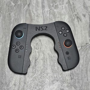 Pode incluir: Um controle de jogo NS2 preto com um design exclusivo em forma de U. Possui dois manípulos analógicos com anéis azuis e laranja, juntamente com botões direcionais e de ação. O controle foi projetado para uma pegada confortável.