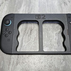 以下が含まれることがあります： Nintendo Switchのロゴが入った黒いゲームコントローラーグリップ。グリップには、両側にボタンとジョイスティックコントロールが付いた2つのハンドルがあります。ジョイスティックには青とオレンジのアクセントが付いています。