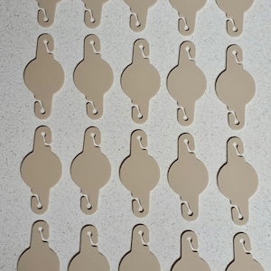 Könnte beinhalten: Darstellung von einundzwanzig beigefarbenen Kleideranhängern. Jeder Aufhänger hat einen abgerundeten oberen Teil mit einem Haken, einen kreisförmigen Körper und eine gebogene Basis. Sie sind in einem Raster angeordnet.