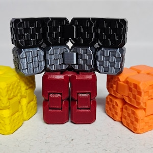 Puede incluir: Cuatro pequeños juguetes en forma de cubo que se entrelazan, de diferentes colores. El juguete de arriba es negro y plateado, el juguete del medio es rojo, el juguete de la izquierda es amarillo y el juguete de la derecha es naranja.