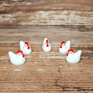 Puede incluir: Cinco pequeñas figuras de gallina de cerámica blanca con crestas y picos rojos están dispuestas sobre una superficie de madera. Las gallinas están en varias poses, algunas mirando hacia adelante y otras giradas hacia un lado. El fondo es de madera marrón cálido.