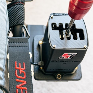 May include: A racing simulator gear shifter with a red handle and a black base. The text "SHH SHIFTER" is visible on the base. The image also shows a person's leg in grey trousers and a black strap.