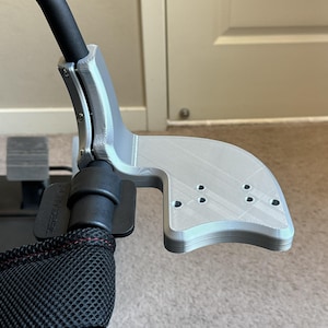 May include: A silver 3D printed racing wheel mount with a black strap and a black mesh fabric. The mount has a curved design and multiple holes.