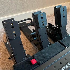 May include: Black racing pedals with a black metal frame and a black strap. The pedals have a black rubber footrest and a black metal base. The pedals are labeled "MOZA RACING".