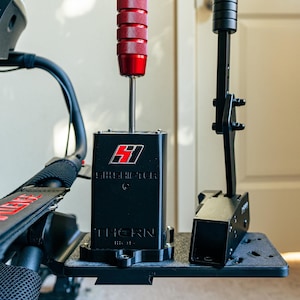 Puede incluir: Un cambio de marchas negro con una perilla roja y el texto "SHIFTER" y "THORN" impreso en él. El cambio de marchas está montado en una placa de metal negro.