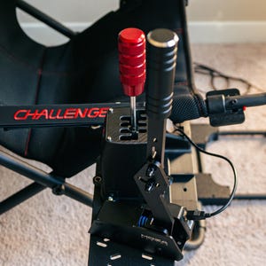 May include: A black shifter with a red knob and a black lever. The shifter is mounted on a black metal plate with the word "MOZA" printed on it. The shifter is connected to a black cable.