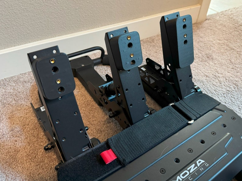 May include: Black racing pedals with a black metal frame and a black strap. The pedals have a black rubber footrest and a black metal base. The pedals are labeled "MOZA RACING".