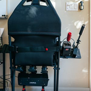 May include: A black Playseat gaming chair with a red shifter mount attached to the side. The chair is folded down, showing the black metal frame and the red release levers. The text "Playseat" is visible on the back of the chair.