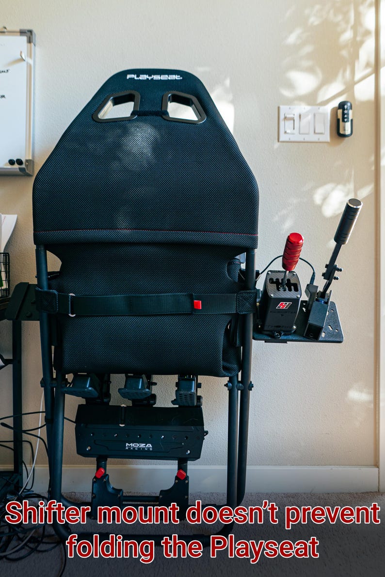 May include: A black Playseat gaming chair with a red shifter mount attached to the side. The chair is folded down, showing the black metal frame and the red release levers. The text "Playseat" is visible on the back of the chair.