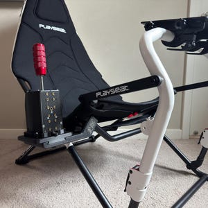 Op de afbeelding: Een zwart-witte Playseat racesimulator stoel met een rode versnellingspook. De stoel heeft een zwarte zitting en een wit frame. De versnellingspook is rood en zilver. Ontworpen voor een realistische race-ervaring.