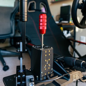 May include: A black racing simulator shifter with a red knob and a black lever. The shifter is mounted on a black metal frame with a USB cable.