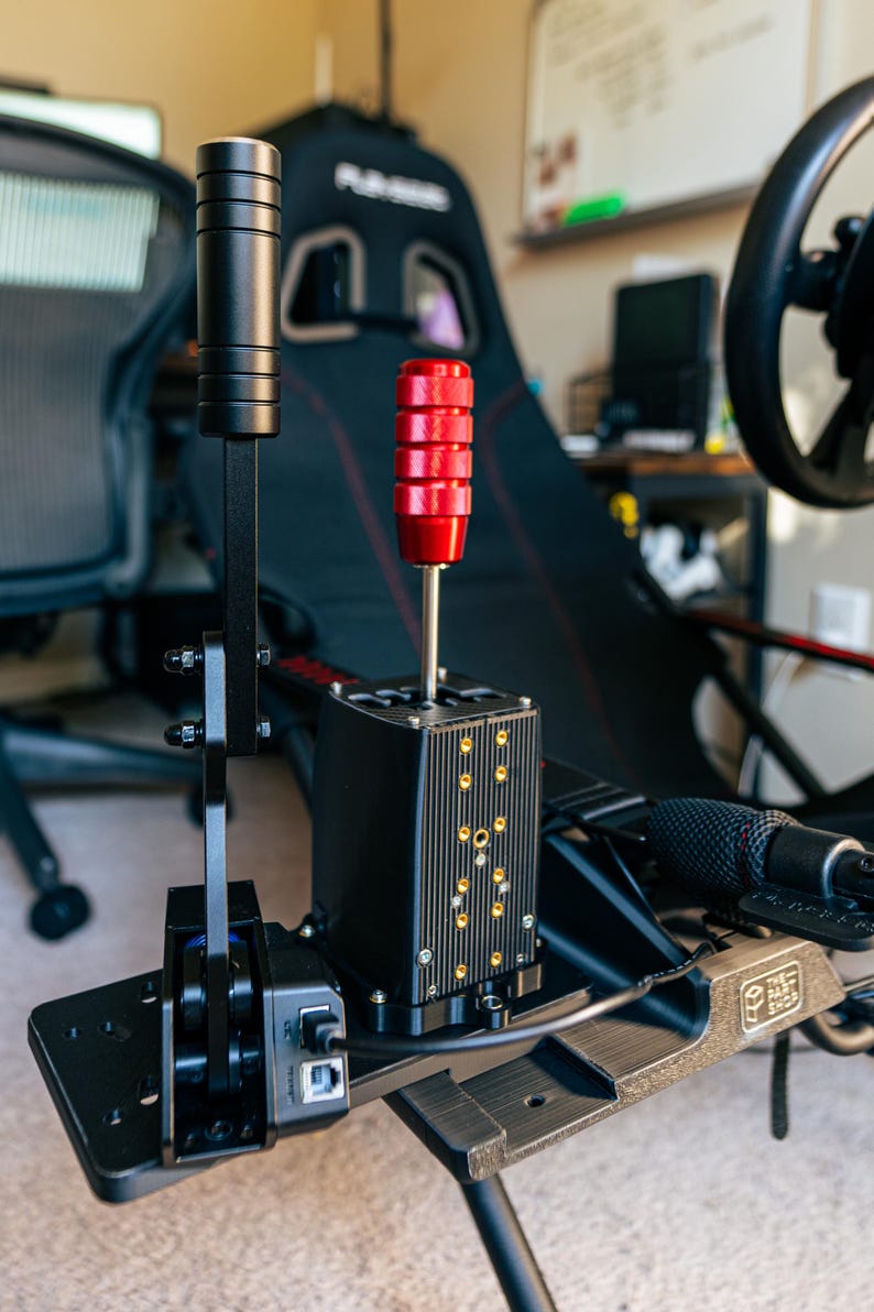 May include: A black racing simulator shifter with a red knob and a black lever. The shifter is mounted on a black metal frame with a USB cable.
